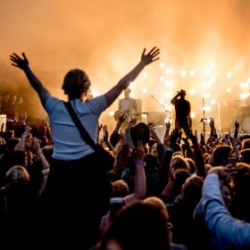 Foran en scene står et band oplyst af orange lys. Publikum står med begge hænder i vejret. En kvinde sidder på skulderen af en anden og hun har begge arme over hovedet. 