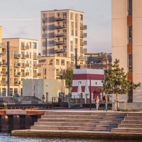 Sollys skinner på lejlighederne ved Odense havn. To mennesker sig en tur ved havnen i det flotte vejr.