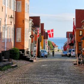 Brostentbelagtgade i Ærøskøbing om sommeren med havkig. Stokroser i forgrunden og farverige huse.