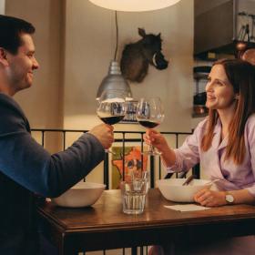 En kvinde og en mand spiser på en restaurant. Der står tallerkener på deres bord og de smiler og skåler i rødvin. 