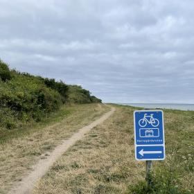 En græseng der grænser op til havet med en lang grussti som fortsætter i horisonten. I forgrunden er et blåt skilt med en cykel, en pil til venstre og navnet Herregårdsruten. 