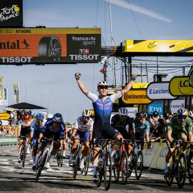 En masse cykelryttere til Tour de France fotograferet ved målområdet. En rytter har armene oppe i sejr.