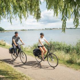 To cyklister med cykeltasker bagpå kører af sted langs vandet.