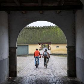 To cyklister ses gennem buet port på brostensbelagt gårdsplads.