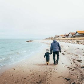 Far og søn har varmt tøj på, og går hånd i hånd på en strand med sommerhuse i baggrunden