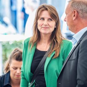 I forgrunden af billedet står en kvinde og mand og snakker sammen. Manden har mørk blazerjakker på og står med siden til. Kvinden har grøn blazer på og kigger på manden der står til højre for hende. Bag dem sidder en kvinde, og i baggrunden står nogle slørede mennesker rundt om et højbord.