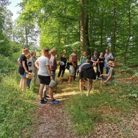 En stor gruppe mennesker står samlet i en cirkel i en grøn skov på en solskinsdag.