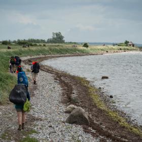 Syv vandrere med hver deres vandrerygsæk går ved strandkanten langs vandet.