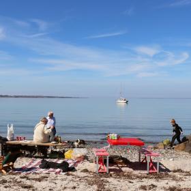 en gruppe af mennesker sidder på en strand med bænke, tæpper, mad og drikke