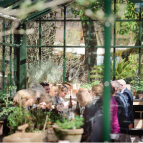 En flok mennesker sider ved et langt bord i en drivhuslignende bygning i grønne omgivelser.