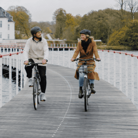 To cyklister cykler på en cykelsti anlagt på vandet med et hvidt gods i baggrunden