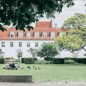 En grøn græsplæne i en park hvor der sidder en gruppe mennesker på et picnictæppe foran et hvidt gods