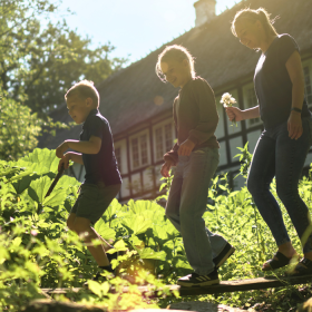To børn og en kvinde går på et bræt midt i et grønt område med et gammeldags bindingsværkshus i baggrunden. 