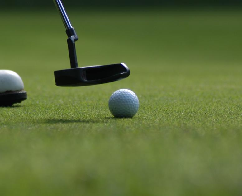 Nærbillede af golfbold på fairway. Golfkølle nærmer sig bolden og det forreste af en hvid golfsko ses til venstre i billedet.