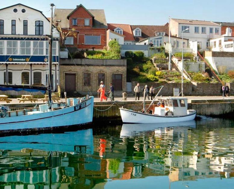 En havn set fra vandet. I vandet er der en lille jolle og en stor gammel sejlbåd. På en bakke over havnen er der placeret gamle bygninger, der har udsigt til havnen.