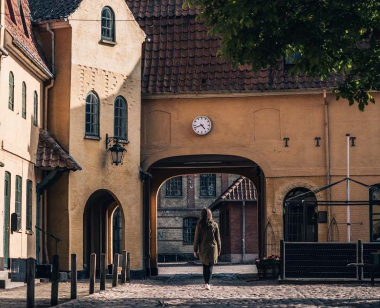 Kvinde på vej ind gennem port i gammel bygning i Assens. Uret over porten viser, at klokken er 17.45.