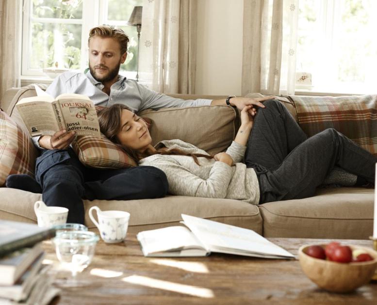 En mand sidder i sofa og læser en bog, imens ligger en kvinde på en pude op ad hans skød og slapper af.