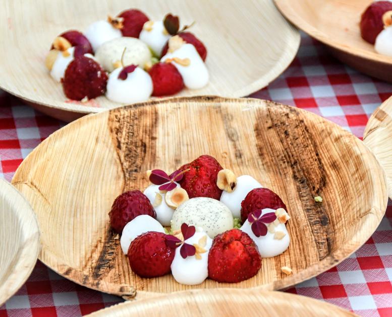 På en rød og hvid ternet dug står otte bæredygtige paptallerkner. På tallerknerne er en flot anrettet dessert bestående af jordbær, hvidt skum og en iskugle i midten. 