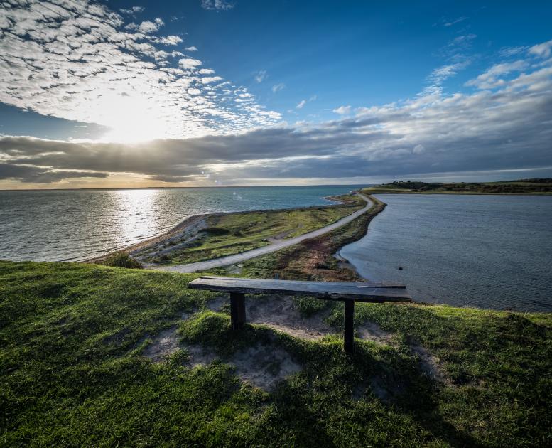 Udsigt over tange med smal vej og vand på begge sider. Solen stråler dramatisk gennem lavthængende grå skyer. I forgrunden står en træbænk.