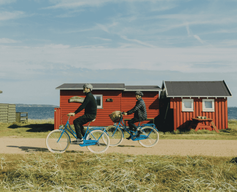 To cyklister kører på en grusvej med græs til den ene side og hav og badehuse til den anden side
