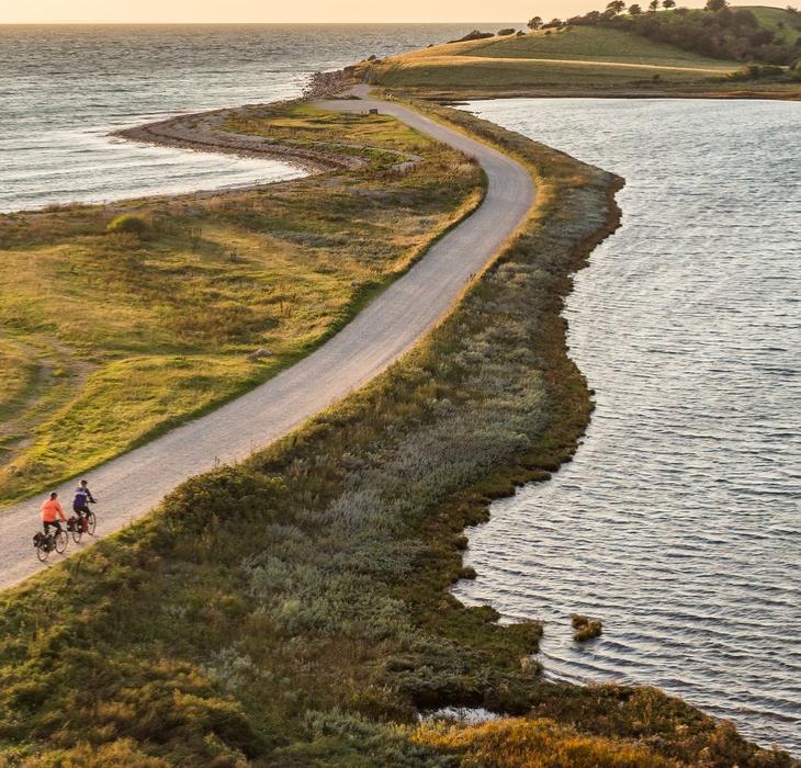 Luftfoto af to cyklister på smal vej ud af tange med vand på begge sider og land i baggrunden.