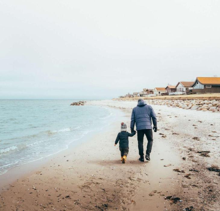 Far og søn har varmt tøj på, og går hånd i hånd på en strand med sommerhuse i baggrunden